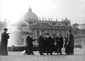 7 Christian Brothers dressed in their robes stand next to a fountain in St. Peter’s Square in Vatican City, with Saint Peter’s Basilica in the background.